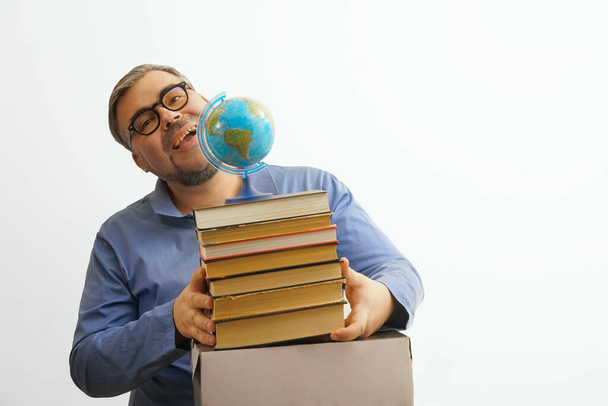 Vousatý šťastný muž s brýlemi na bílém pozadí drží glóbus v ruce, zatímco drží hromadu knih - Fotografie, Obrázek