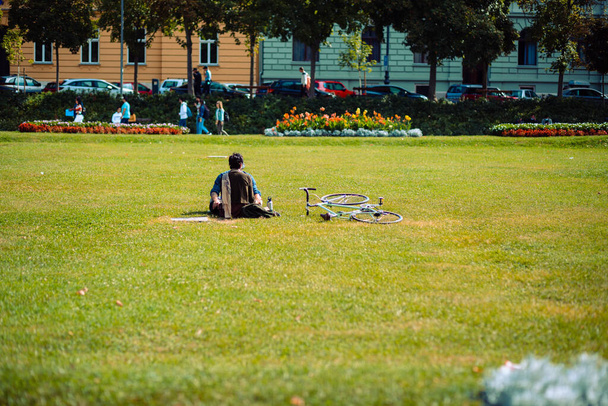A back view of a male sitting on the green grass and relaxing near his bicycle in on of  Zagreb parks - Фото, изображение