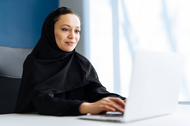Beautiful woman with abaya dress working on her computer. Middle aged female employee at work in a business office in Dubai. Concept about middle eastern cultures and lifestyle - Foto, afbeelding
