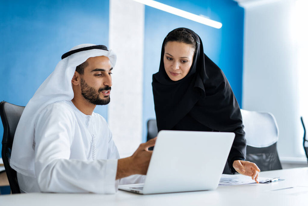 Man and woman with traditional clothes working in a business office of Dubai. Portraits of  successful entrepreneurs businessman and businesswoman in formal emirates outfits. Concept about middle eastern cultures, lifestyle and professional occupatio - Φωτογραφία, εικόνα