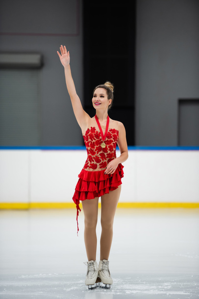 happy figure skater in red dress and golden medal waving hand on ice arena - Foto, Bild