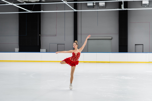 volledige lengte van kunstschaatser in rode jurk het uitvoeren van kameel spin in ijs arena - Foto, afbeelding