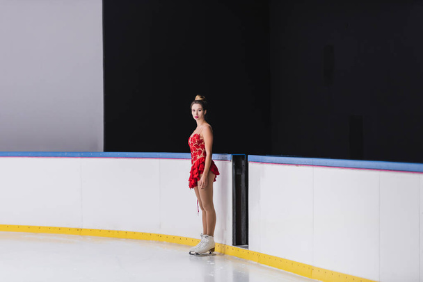 pełna długość łyżwiarki figurowej w czerwonej sukience stojącej i patrzącej w kamerę na lodowisku  - Zdjęcie, obraz