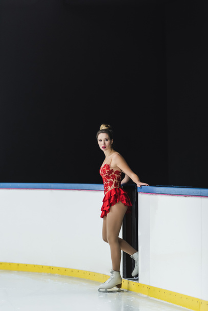 longitud completa de joven preocupado patinador artístico en vestido rojo de pie cerca de la arena de hielo - Foto, Imagen
