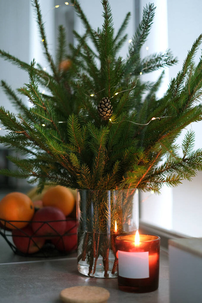Las ramas de coníferas de un árbol de Navidad están en un jarrón transparente con velas sobre la mesa. ramo de Navidad en la casa - Foto, imagen