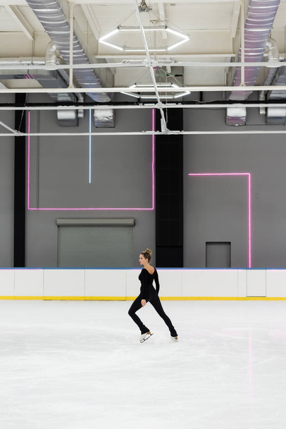 full length of young woman in black bodysuit skating in professional ice arena - Photo, Image