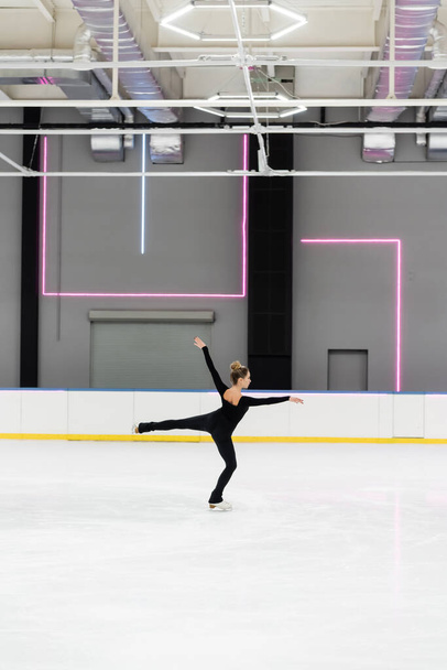 volledige lengte van jonge vrouw in zwart bodysuit met uitgestrekte handen figuur schaatsen in professionele ijs arena - Foto, afbeelding