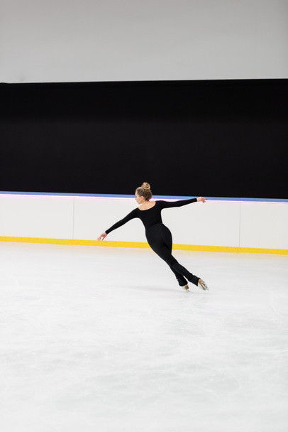 volledige lengte van professionele kunstschaatser in zwart bodysuit schaatsen met uitgestrekte handen in de ijshal - Foto, afbeelding