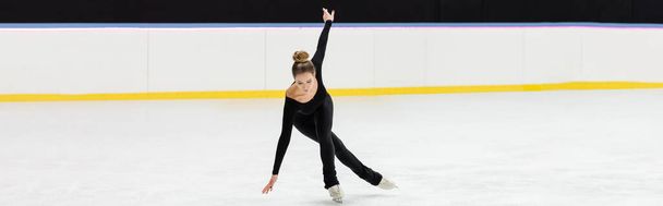 full length of professional figure skater in black bodysuit skating with outstretched hands in ice arena, banner - Photo, Image