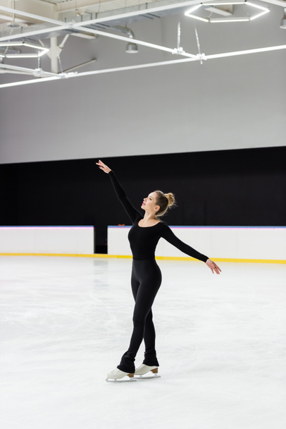 pełna długość zmysłowej łyżwiarki figurowej w czarnym body skating z wyciągniętymi rękami na arenie lodowej  - Zdjęcie, obraz