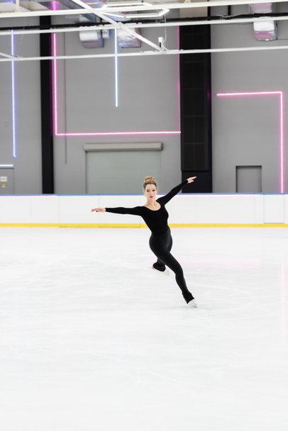 full length of professional figure skater in black bodysuit skating with outstretched hands in ice arena with neon lights - 写真・画像