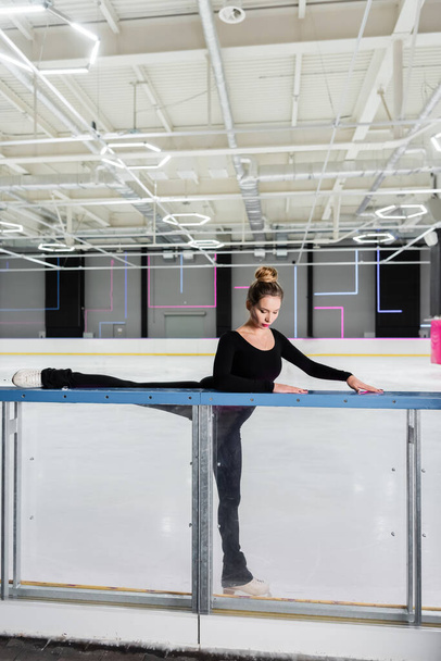 young woman in white figure skates stretching near ice arena - Zdjęcie, obraz