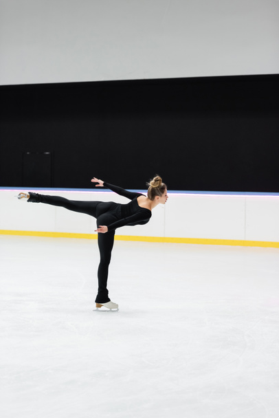 side view of professional figure skater in black bodysuit skating with outstretched hand in ice rink - Foto, Bild