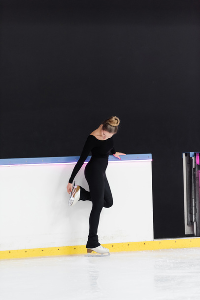 full length of young figure skater in black bodysuit checking blade on ice skates near frozen ice arena - Photo, Image