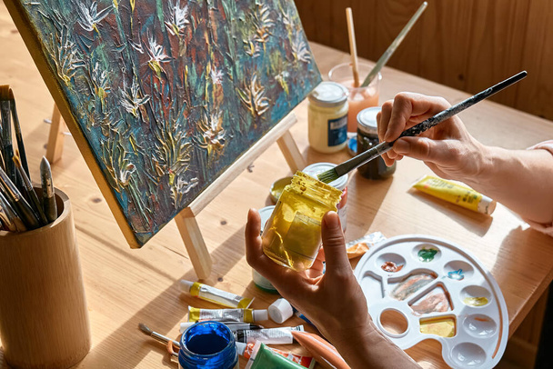 Mujer tomando un color con pincel para pintar sobre lienzo en el caballete cerca de la ventana en el estudio. Pasatiempo de pintura artística. Ocio creativo. Concepto de auto expresión. - Foto, Imagen