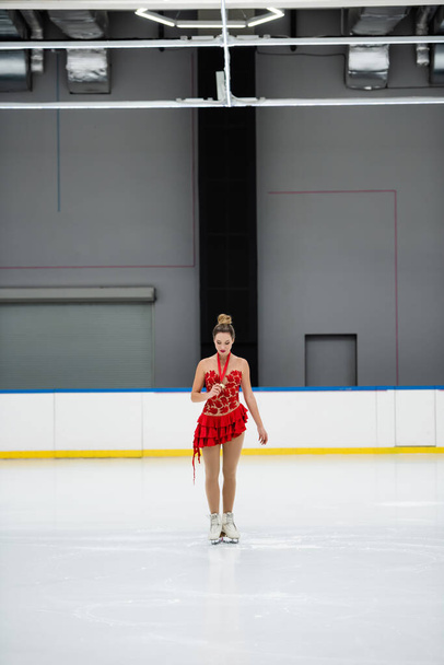 volledige lengte van de jonge kunstschaatser in rode jurk kijken naar gouden medaille op de ijsbaan - Foto, afbeelding