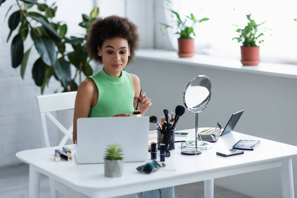 Elinde kozmetik fırçası olan Afrikalı Amerikalı bir kadın ve evinde kozmetik ürünlerinin yanındaki laptopa bakıyor.  - Fotoğraf, Görsel