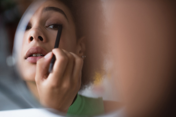 Mujer afroamericana aplicando delineador de ojos cerca del espejo  - Foto, imagen