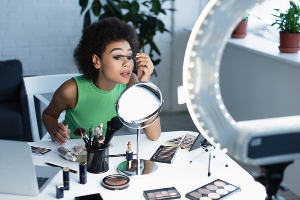 Pěkný africký americký blogger aplikující řasenku v blízkosti zrcadla, gadgets a dekorativní kosmetiky doma  - Fotografie, Obrázek