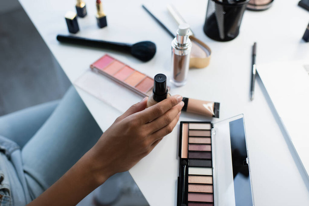Cropped view of african american woman holding face foundation near blurred cosmetics on table  - Photo, Image