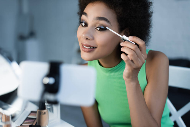 Positive african american blogger looking at smartphone while applying eye shadows  - Photo, Image