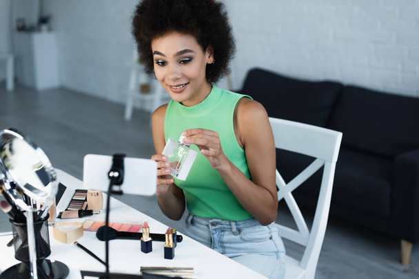 Neşeli Afro-Amerikan blog yazarı kozmetik ve akıllı telefonun yanında parfüm tutuyor.  - Fotoğraf, Görsel