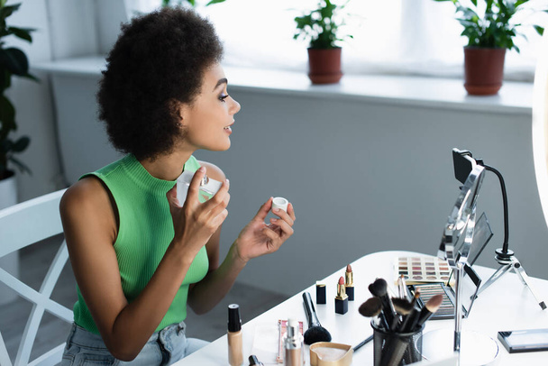Zijaanzicht van Afrikaanse Amerikaanse blogger spuitende parfum in de buurt van cosmetica en smartphone thuis  - Foto, afbeelding