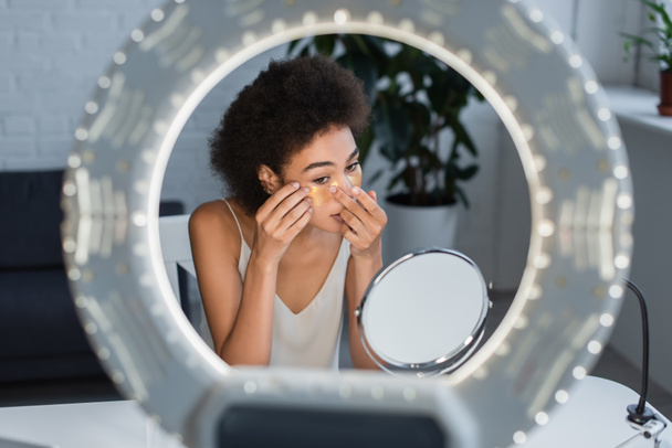 Young african american woman applying eye patch near mirror and ring light at home  - Photo, Image