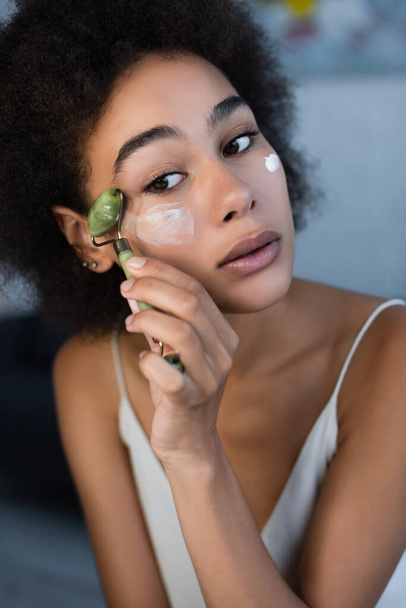 Jeune femme afro-américaine appliquant crème cosmétique avec rouleau de jade à la maison  - Photo, image