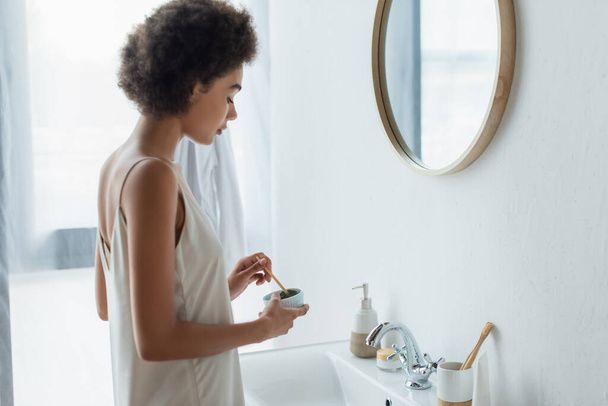 Boční pohled na mladé africké Američanky míchání obličeje maska v blízkosti zrcadla v koupelně  - Fotografie, Obrázek