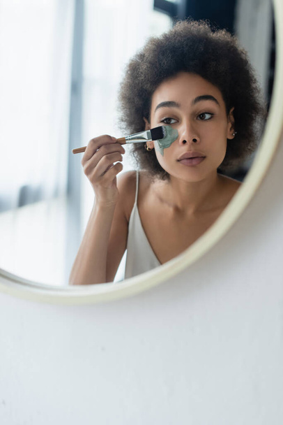 Mujer afroamericana aplicando mascarilla con cepillo cosmético cerca del espejo en casa  - Foto, Imagen