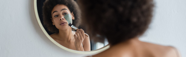 Mujer joven afroamericana con cepillo cosmético aplicando mascarilla cerca del espejo, pancarta  - Foto, imagen