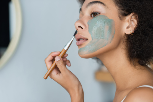Genç Afrikalı Amerikalı kadın banyoda yüzüne kilden maske takıyor.  - Fotoğraf, Görsel