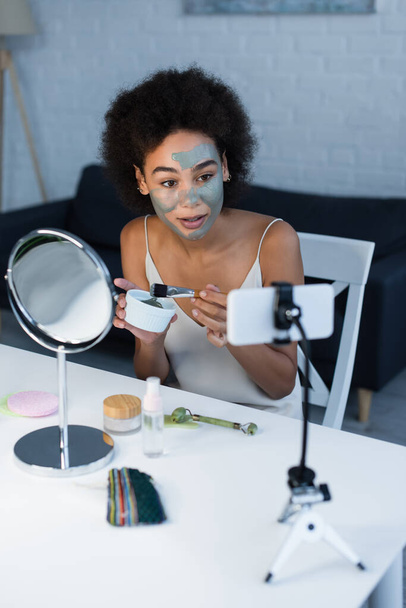 blogger afroamericano sosteniendo máscara de arcilla y cepillo cosmético cerca de teléfono inteligente en casa  - Foto, Imagen