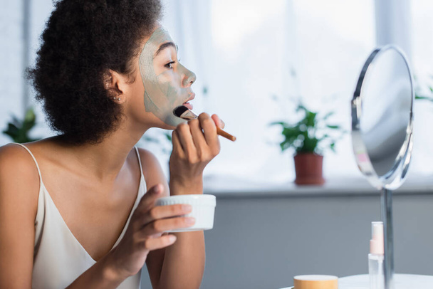 自宅で鏡の近くに顔マスクを適用アフリカ系アメリカ人女性の側面図  - 写真・画像