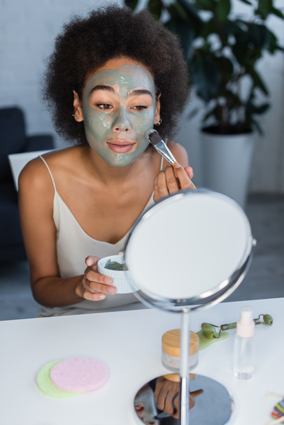 Mujer afroamericana aplicando mascarilla cerca de espejo y rodillo de jade en casa  - Foto, Imagen