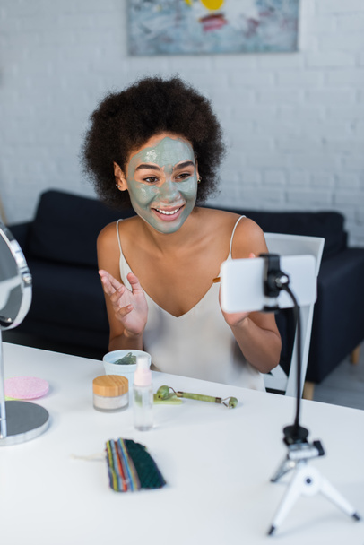 Smiling african american blogger with face mask looking at smartphone at home  - 写真・画像