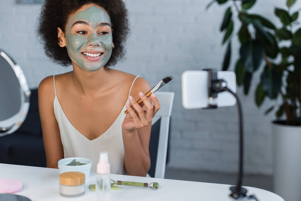 Mujer afroamericana sonriente con máscara de arcilla sosteniendo cepillo cosmético cerca del teléfono celular en casa  - Foto, imagen