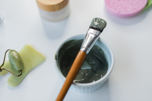 Bovenaanzicht van cosmetische borstel op kleimasker bij jade roller en cosmetische crème op tafel  - Foto, afbeelding