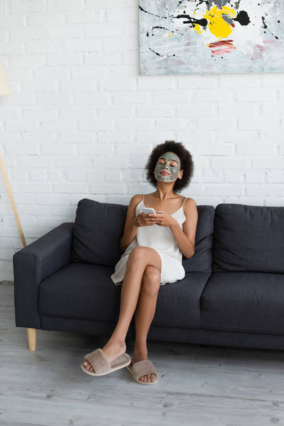Young african american woman with clay mask on face using smartphone on couch at home  - Photo, image