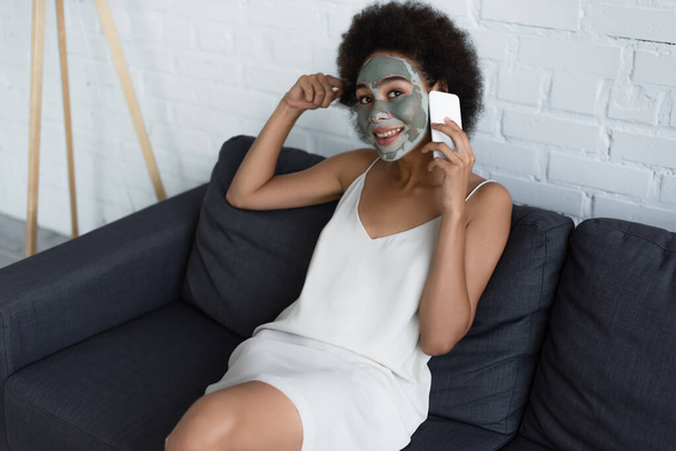 Smiling african american woman with clay mask on face talking on smartphone on couch  - Foto, Imagem