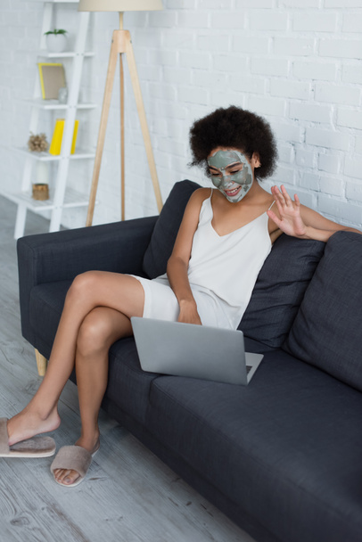 Улыбающаяся африканская американка в глиняной маске на лице с видеозвонком на домашний ноутбук  - Фото, изображение