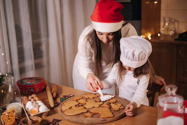 Mãe em chapéu de santas com seu pouco mais doce decora pão de gengibre usando esmalte. Conceito de tradições de Natal e Ano Novo. Padaria de Natal. Boas festas - Foto, Imagem