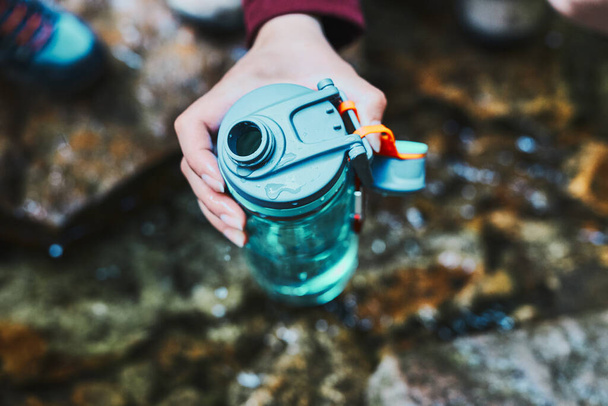 Woman taking pure water to bottle from mountain stream during trekking in mountains. Hiker crouching on rocks, filling bottle up with cold mountain water. Enjoying the outdoors in the summer trip vacation - Foto, afbeelding