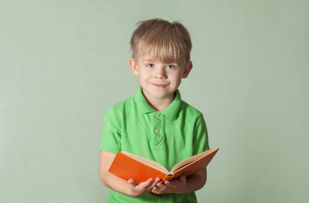 a blond boy in a green T-shirt with a smile stands with a book. green background. - Foto, imagen