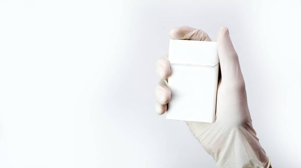 Una mano in un guanto medico tiene un pacchetto bianco di sigarette, un concetto anti-fumo. - Foto, immagini