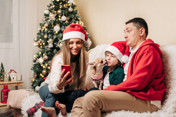 Famille heureuse avec fils enfant ayant chat vidéo au téléphone. Mère dans le chapeau de Père Noël, père, enfant garçon et chien en pull s'amuser pendant les vacances de Noël à la maison. Joyeux Noël et bonne fête de l'année - Photo, image