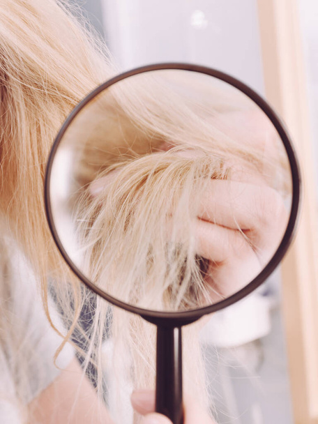 Haircare, bad effects of bleaching concept. Woman looking at ends of her blonde hair through magnifying glass - Foto, Imagen