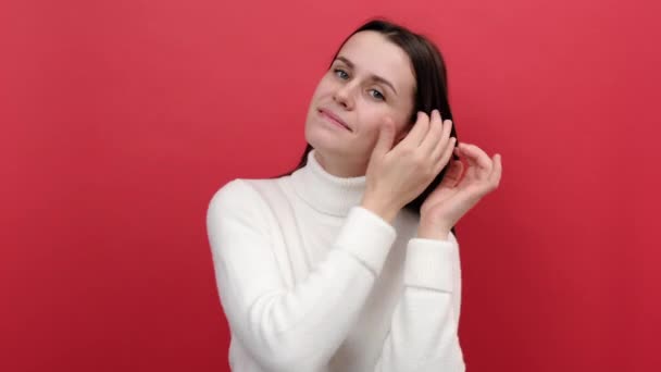 Portrait of cute attractive young woman looking in mirror, admiring herself, combing her hair, doing care procedures, dressed in white knitted sweater, posing isolated on red studio background wall - Felvétel, videó