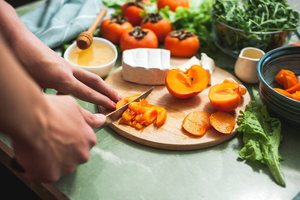 Ingredients and process of making a vitamin winter salad with persimmons, tangerines and cheese Female or teenage hand cuts persimmon on a round wooden board Vegan lunch preparation. Selective focus - Фото, изображение